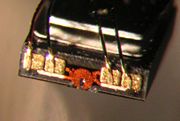Microphotograph of a hard disk head. The size of the front face  is about 0.3 mm × 1.0 mm. The (not visible) bottom face of the slider is about 1.0 mm × 1.25 mm and faces the platter. One functional part of the head is the round, orange structure in the middle—the lithographically defined copper coil of the write transducer. Also note the electric connections by wires bonded to gold-plated pads.