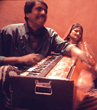 Man playing a harmonium.  He is pumping the bellows of the harmonium with one hand and playing the keys with the other.