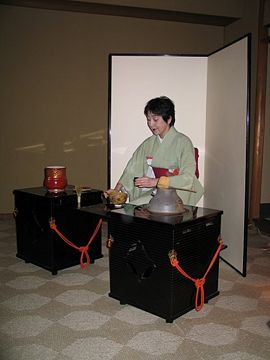 A woman wearing kimono and seated at a tana performs the Ryū-rei ceremony. Visible from far left to right are the red fresh water container (its lid is placed on the tana), the tea whisk, the tea bowl, the iron kama (pot) and the ladle (resting on the pot). The woman is holding the natsume.