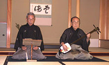 A Japanese man playing a shamisen while another sings.