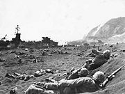 Marines burrow in the volcanic sand on the beach of Iwo Jima, as their comrades unload supplies and equipment from landing vessels despite the heavy rain of artillery fire from enemy positions in the background